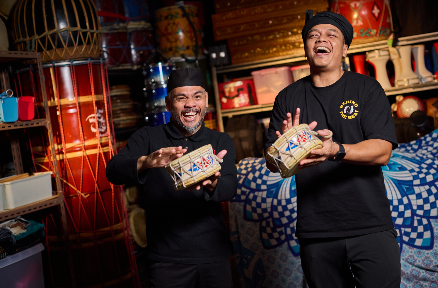 Enter a descriptive altTwo men in black outfits laugh joyfully while holding traditional drums, surrounded by a collection of traditional instruments in the background text.